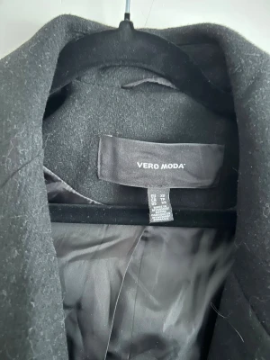 Svart kappa från Vero Moda - Stilren svart kappa från Vero Moda med knytskärp i midjan och breda slag. Kappan är helfodrad och har en klassisk, lång siluett som passar perfekt till höst och vinter. Materialet är mjukt och ger en snygg look till alla outfits.