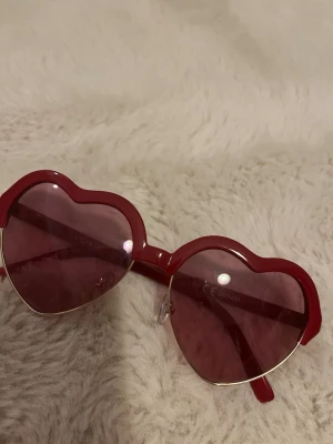 Röda hjärtformade solglasögon - Säljer ett par riktigt snygga solglasögon med hjärtformade bågar i rött och matchande röda glas. Perfekta för dig som vill sticka ut och addera en lekfull touch till din stil. Glasögonen har näskuddar för extra komfort.