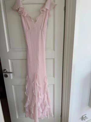Ljusrosa maxiklänning med volangdetaljer - Ljusrosa maxiklänning med volanger längs axlar och kjol. Klänningen är aldrig använd och har prislapp kvar. Klänningen har v-ringning både fram och bak och formar kroppen på ett fint sätt. Perfekt för speciella tillfällen som bal och bröllop. 