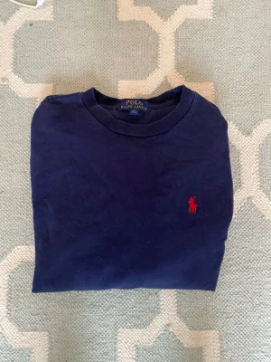Mörkblå långärmad tröja Polo Ralph Lauren - Mörkblå långärmad tröja från Polo Ralph Lauren med klassisk rund halsringning och röd broderad logga på bröstet. Tillverkad i mjuk bomull som känns skön mot huden. Perfekt för en avslappnad och stilren look.