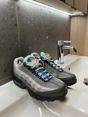 Nike Air Max 95 grå/blå sneakers - Nike Air Max 95 sneakers i grått och svart med blå detaljer. Ovandelen är i mesh och mocka med vita snören och synlig Air Max-dämpning i sulan. Klassisk vågig design och retrovibe. Perfekta för dig som gillar streetwear och sportig stil.