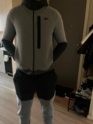 Nike grå tracksuit med huva - Nike tracksuit i mjukt material, ljusgrå med svarta detaljer. Jackan har hel dragkedja, huva och en vertikal bröstficka. Byxorna har svarta partier vid fickorna och knäna. Perfekt för chill eller träning.