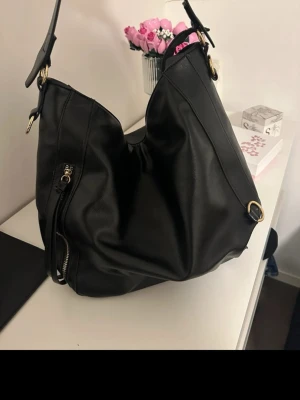 Svart axelremsväska i skinnimitation - Stilren svart handväska i mjuk skinnimitation med axelrem och guldfärgade metalldetaljer. Väskan har en praktisk dragkedjeficka på framsidan och rymligt huvudfack. Perfekt för dig som vill ha en enkel men snygg accessoar till din outfit.