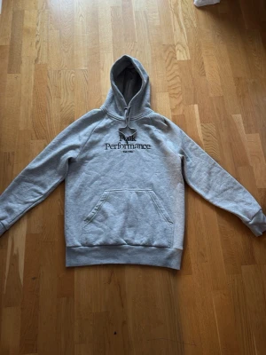 Grå hoodie från Peak Performance - Snygg grå hoodie från Peak Performance med svart logga på bröstet. Klassisk känguruficka framtill och justerbar huva med dragsko. Tillverkad i mjukt material som passar perfekt för en avslappnad stil.