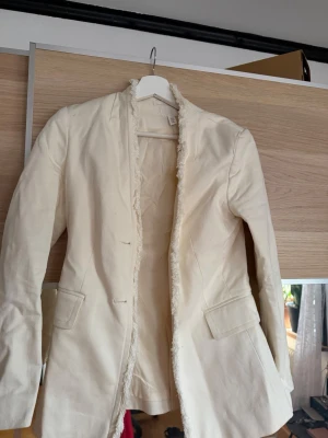 Krämvit kavaj med råa kanter H&M - Säljer en krämvit kavaj från H&M i storlek 34. Kavajen har råa, fransiga kanter längs öppningen och två fickor framtill. Materialet är kraftigt och ger en cool, avslappnad look. Perfekt för dig som vill ha en unik twist på din outfit.