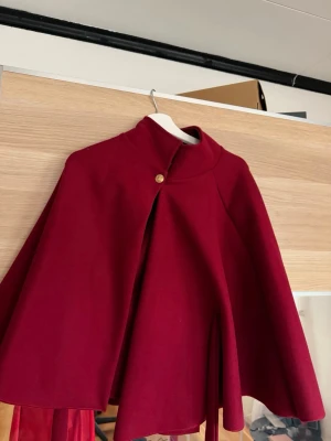 Vinröd cape-kappa med knapp - Stilren vinröd cape-kappa med hög krage och en dekorativ guldig knapp vid halsen. Kappan har en vid, svepande siluett och är tillverkad i ett tjockare tyg som ger ett elegant intryck. Perfekt för dig som vill sticka ut med en unik och trendig look.