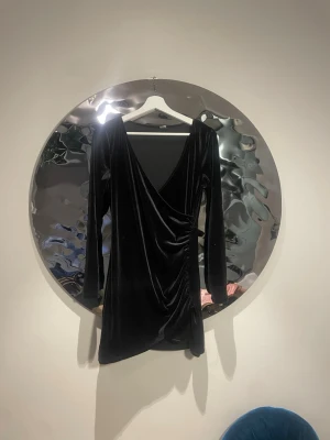 Svart omlottklänning i sammet - Snygg svart kortklän clouding i mjuk sammet med omlottdesign och djup v-ringning. Klänningen har långa ärmar och draperad detalj i sidan som ger en elegant siluett. Perfekt för dig som vill sticka ut med stil.