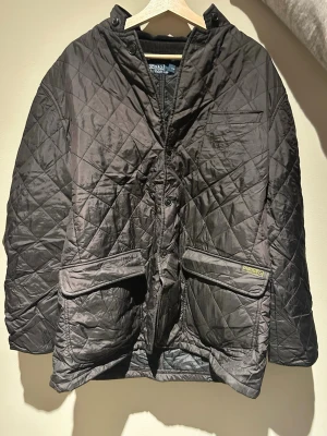Svart quiltad vindjacka Polo Ralph Lauren - Quiltad svart vindjacka från Polo Ralph Lauren med två stora fickor framtill, bröstficka och knäppning. Jackan har en krage i manchestertyg och diskret logga på fickan. Perfekt för dig som gillar klassisk och stilren design.