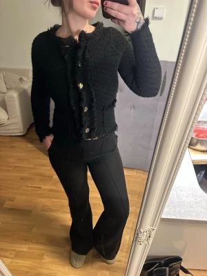 Svart fransig kofta med gulddetaljer - Trendig svart kofta med fransiga kanter och dekorativa guldfärgade knappar framtill. Kortare modell med lång ärm och strukturerat tyg som ger en cool look. Perfekt att styla till både jeans och kjol för en chic outfit.