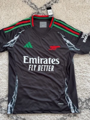 Arsenal svart matchtröja Adidas - Snygg svart Arsenal matchtröja från Adidas med gröna och röda detaljer på axlarna, klubbmärke i rött och sponsortryck framtill. Tillverkad i lätt och ventilerande material, perfekt för träning eller match. Kortärmad med V-ringning och grafiska mönster på sidorna.