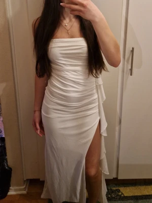 Vit långklänning med slits - Säljer en elegant vit långklänning med axelbandslös design och hög slits på sidan. Klänningen har draperade detaljer längs midjan och en figurnära passform som framhäver siluetten. Perfekt för dig som vill sticka ut med en stilren look.