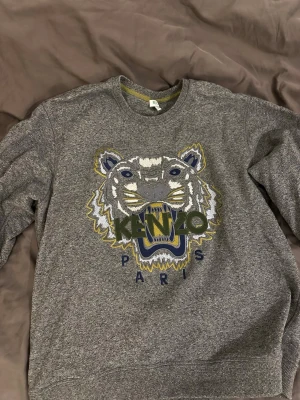 Grå Kenzo sweatshirt med tiger - Säljer en grå sweatshirt från Kenzo med det ikoniska tigerhuvudet broderat framtill i vitt, blått, gult och grönt. Tröjan har rund halsringning och ribbade muddar. Perfekt för dig som vill ha en statement piece från ett känt märke.
