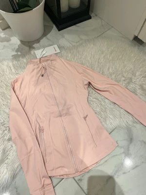 Ljusrosa träningsjacka från Lululemon - Snygg ljusrosa träningsjacka från Lululemon med hel dragkedja framtill och två fickor med dragkedja. Jackan har hög krage och figurnära passform, perfekt för löpning eller gym. Tillverkad i ett stretchigt funktionsmaterial som andas.