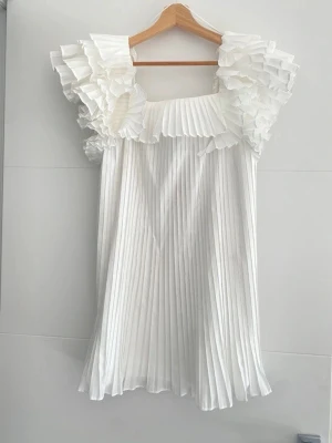 Zara Vit plisserad kortklänning - Supersöt vit kortklänning med plisserat tyg och volangdetaljer vid axlarna. Klänningen har en luftig passform och är ärmlös, perfekt för sommarens alla tillfällen. Ger en romantisk och trendig vibe.