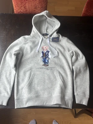 Grå hoodie Polo Bear Ralph Lauren - Helt oanvänd, storlek M men sitter som S på mig som är 183cm. Säljer pågrund av att den var lite för liten för min smak. Sitter nog perfekt på dig som är strax under 180 och gillar lite mer slim hoodies.