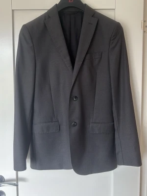 Mörkgrå kavaj från J.Lindeberg - Stilren mörkgrå kavaj från J.Lindeberg i slim fit-modell. Klassisk design med två knappar, smala slag och tre fickor framtill. Tillverkad i ett slätt tyg som ger en modern och snygg look. Perfekt för dig som gillar en skräddad siluett. Endast använd en gång och är perfekt för dig som slutar skolan till sommaren eller som ska på ett speciellt event. Storlek 46 slim fit