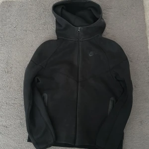Svart Nike tech hoodie med dragkedja - Säljer en svart hoodie från Nike med hel dragkedja framtill och två fickor med dragkedja. Hoodien har en hög krage och en stor huva. Tillverkad i mjukt material som känns skönt mot huden. Diskret Nike-logga på bröstet för en clean look.