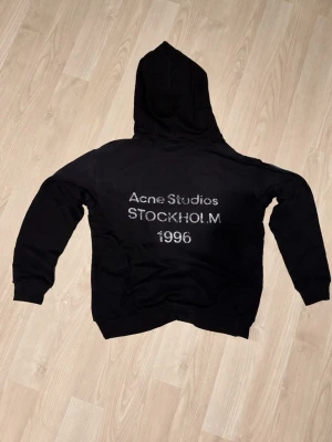 Svart hoodie Acne Studios Stockholm - Svart hoodie från Acne Studios med tryck på ryggen där det står 'Acne Studios STOCKHOLM 1996'. Klassisk passform med huva och ribbade muddar. Perfekt för en avslappnad och trendig look. Materialet känns mjukt och bekvämt. Kvitto finns! 