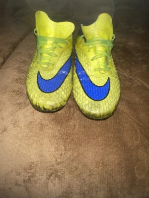 Gula Nike fotbollsskor med blå swoosh - Säljer ett par gula Nike fotbollsskor med stor blå swoosh på sidan. Skorna har gröna skosnören och en texturerad ovandel för bättre bollkontroll. Yttersulan har dobbar som passar för spel på gräs. Perfekta för dig som vill sticka ut på planen.