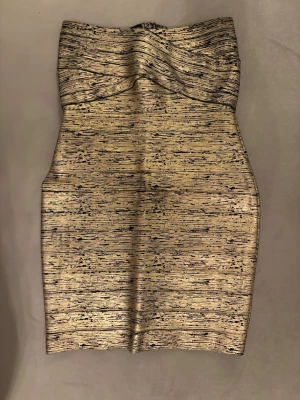 Guldig axelbandslös bandage klänning - Snygg axelbandslös kort klänning i guld med svart mönster. Klänningen har en figurnära passform och dragkedja baktill för enkel påklädning. Materialet är glittrigt och ger en lyxig känsla, perfekt för att sticka ut.