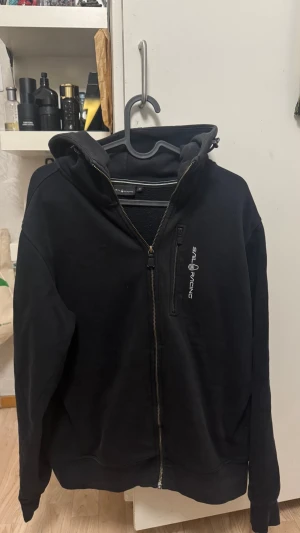 Svart hoodie från Sail Racing - Säljer en svart hoodie från Sail Racing med dragkedja framtill och en bröstficka med logga. Hoodien har en stor huva och ribbade muddar vid ärmar och nederkant. Perfekt för dig som gillar en clean och sportig stil. Materialet känns mjukt och bekvämt. Säg till om du behöver fler bilder 🙌
