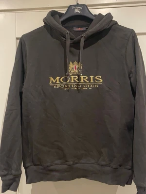 Mörkgrå hoodie från Morris - Snygg mörkgrå hoodie från Morris med broderad logga och text 'Morris Sporting Club' i guld på bröstet. Klassisk huva med dragsko och ribbade muddar. Perfekt för en chill och stilren look.