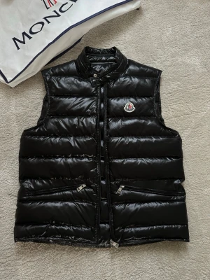 Svart dunväst från Moncler - Snygg svart dunväst från Moncler med glansig finish och klassisk logga på bröstet. Västen har ståkrage, två dragkedjefickor och quiltad design. Perfekt för lager-på-lager och streetstyle.