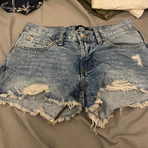 Blå denimshorts med slitningar - Säljer ett par blå denimshorts från 157 med råa, fransiga kanter och slitningar på framsidan. Klassisk femficksdesign och knappstängning. Perfekta för sommaren.