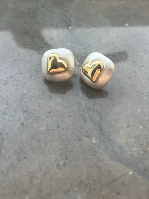 Pärlörhängen med guldfärgade hjärtan - Säljer ett par unika örhängen med oregelbundna pärlliknande former och guldfärgade hjärtan i mitten. Örhängena har en cool och trendig look som sticker ut. Perfekta för dig som gillar statement-smycken och vill ha något annorlunda.