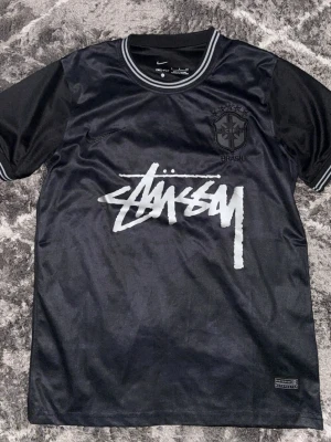 Svart Stüssy x Nike Brasil T-shirt - Snygg svart t-shirt från Stüssy och Nike med Brasil-märke på bröstet och stor vit Stüssy-logga framtill. Tillverkad i ett sportigt, glansigt material med rund hals och korta ärmar. Perfekt för dig som gillar streetwear och unika samarbeten.