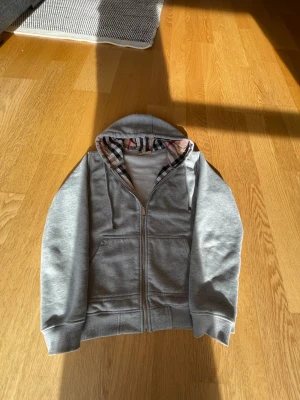 Grå hoodie med dragkedja från Burberry - Snygg grå hoodie från Burberry med dragkedja och klassiskt rutigt foder i huvan. Hoodien har två fickor fram och diskret Burberry-logga broderad på bröstet. Tillverkad i mjukt bomullsmaterial och har ribbade muddar vid ärmar och nederkant.