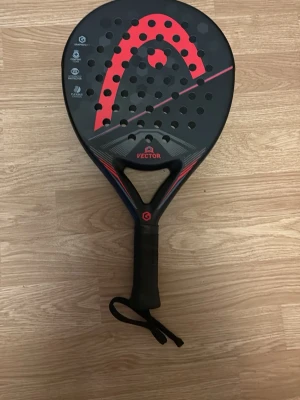 HEAD Vector padelracket svart/röd - Säljer ett HEAD Vector padelracket i svart med röda detaljer. Racketen har rund form, perforerad träffyta och handledsrem. Tillverkat i slitstarkt material och har en modern design med tydlig HEAD-logga på sidan.