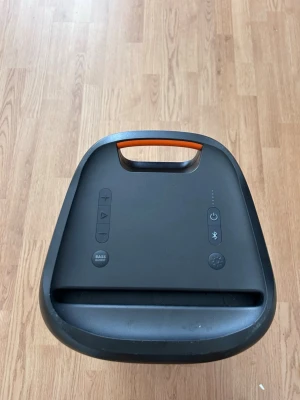 JBL PartyBox 100 - Kraftfull trådlös högtalare från JBL i svart med orange detaljer. Robust design med bärhandtag, tydliga knappar för volym, basförstärkning och Bluetooth. Perfekt för att ta med sig musiken överallt. JBL-logga på sidan och slitstarkt material.
