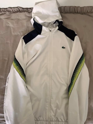 Vit vindjacka från Lacoste med huva - Snygg vit vindjacka från Lacoste med huva och dragkedja. Jackan har svarta detaljer på axlarna och coola gula och svarta mönster längs ärmarna. Klassisk Lacoste-logga på bröstet och meshfoder på insidan för extra komfort.