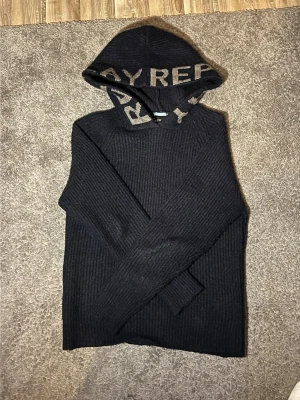 Mörkblå stickad hoodie från Replay - Snygg mörkblå stickad hoodie från Replay med ribbad struktur och stor huva. Huvan har beige textdetaljer som ger en cool kontrast. Perfekt för dig som vill ha en avslappnad men trendig look. Tillverkad i mjukt material för extra komfort.