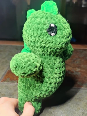 Grön virkad dinosaurie mjukisdjur - Supergullig grön virkad dinosaurie i mjukt garn med svarta och vita ögon. Den har små taggar längs ryggen och en rundad kropp. Perfekt som dekoration eller present till någon som gillar söta figurer.