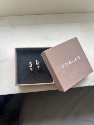 Silvriga örhängen från Edblad - Säljer ett par stilrena silvriga örhängen från Edblad. Örhängena har en modern, avlång form med små nitar längs framsidan som ger en cool och trendig look. Perfekta för dig som gillar minimalistiska accessoarer i metall.