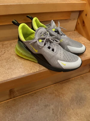 Nike Air Max 270 grå/gul sneakers - Nike Air Max 270 sneakers i grå mesh med neongula detaljer och svart sula. Skorna har en synlig Air-enhet i hälen, ventilerande ovandel och klassisk Nike-logga på sidan. Snygg design med snörning och bekväm passform. Perfekta för dig som gillar streetwear.