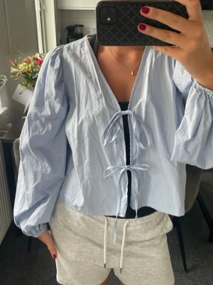 Ljusblå blus med knyt från Gina Tricot - Supersöt ljusblå blus från Gina Tricot med två knyt framtill och puffiga långärmar. Blusen har en avslappnad passform och är gjord i ett tunt, krispigt bomullsliknande material. Perfekt att styla över ett linne eller topp för en trendig look.