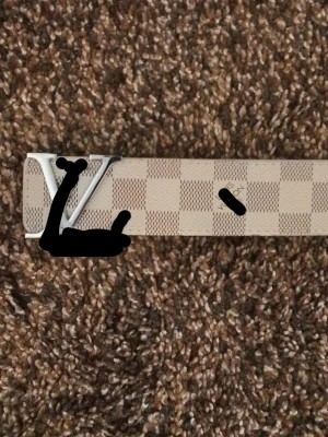 Beige rutigt IV-bälte i skinn - Snyggt beige bälte från med klassiskt rutigt mönster och stor silvrig IV-logga som spänne. Bältet är gjort i skinn och har en stilren design som passar till många olika outfits.