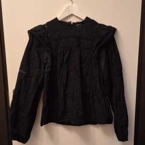 Svart blus med spets från Vintage Stories - Svart blus från Vintage Stories med fina spetsdetaljer vid axlar och ärmar. Blusen har rynkade partier upptill och en luftig passform.