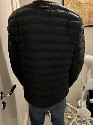 Svart pufferjacka från GANT - Snygg svart pufferjacka från GANT med diskret logga på bröstet. Jackan har quiltad design, dragkedja framtill och ribbade muddar vid halsen. Perfekt för kalla dagar och enkel att matcha med olika outfits.  Köpte den sedan insåg jag att den var för stor darför säljer jag den. 