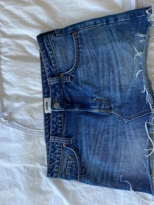 Blå jeansshorts från Monki - Säljer ett par klassiska blå jeansshorts från Monki med rå kant och femficksmodell. Shortsen har hög midja och en avslappnad passform. Perfekta för sommardagar och festivaler.