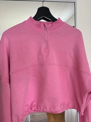 Rosa cropped hoodie från G- Tech - Säljer en snygg rosa cropped hoodie från G Tech med halv dragkedja framtill och broderad logga på bröstet. Tröjan har långa ärmar, ribbad krage och dragsko i nederkanten. Perfekt för en chill och trendig look.
