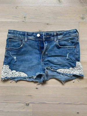 Blå jeansshorts med spetsdetaljer - Säljer ett par blåa jeansshorts med detaljer nedtill. De är coola med lite slitage här och där. Jag har aldrig använt dem då de var för små redan från början. 