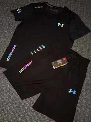 Svart träningsset från Under Armour - Snyggt svart träningsset från Under Armour med t-shirt och shorts. Båda plaggen har regnbågsfärgade loggor och detaljer. Tillverkade i ett lätt och snabbtorkande material som passar perfekt till gymmet eller löpning. Modern och sportig look.