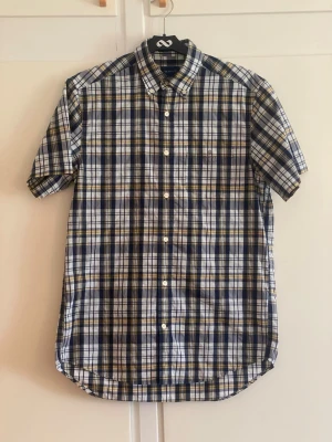 Rutig kortärmad skjorta från GANT - Kortärmad skjorta från GANT i regular fit med rutigt mönster i blått, vitt och gult. Skjortan har klassisk krage, knappar framtill och bröstficka. Tillverkad i ett lätt och bekvämt bomullsmaterial, perfekt för en avslappnad och stilren look.