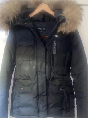 Svart dunjacka med pälskantad huva - Snygg svart dunjacka från Hollies med stor pälskantad huva och flera fickor framtill. Jackan har dragkedja och knappar, samt en figurnära passform. Perfekt för kalla vinterdagar och riktigt varm tack vare dunfyllningen.                                Ny pris 4000kr