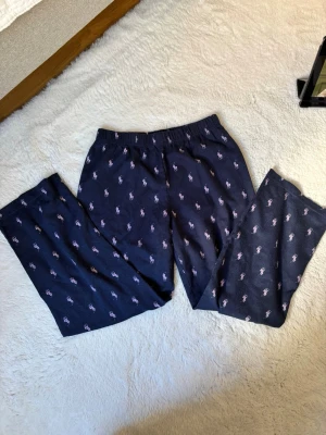 Mörkblå pyjamasbyxor Polo Ralph Lauren - Mörkblå pyjamasbyxor från Polo Ralph Lauren med elastisk midja och ett mönster av små rosa och vita loggor. Tillverkade i mjukt tyg som passar perfekt för avslappnade kvällar hemma. Klassisk och stilren design.