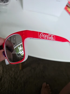 Röda Coca-Cola solglasögon - Snygga solglasögon med röd och vit båge och spegelglas. Klassisk fyrkantig form och Coca-Cola-logga på sidan. Perfekta för dig som vill sticka ut med en ikonisk accessoar i sommar.
